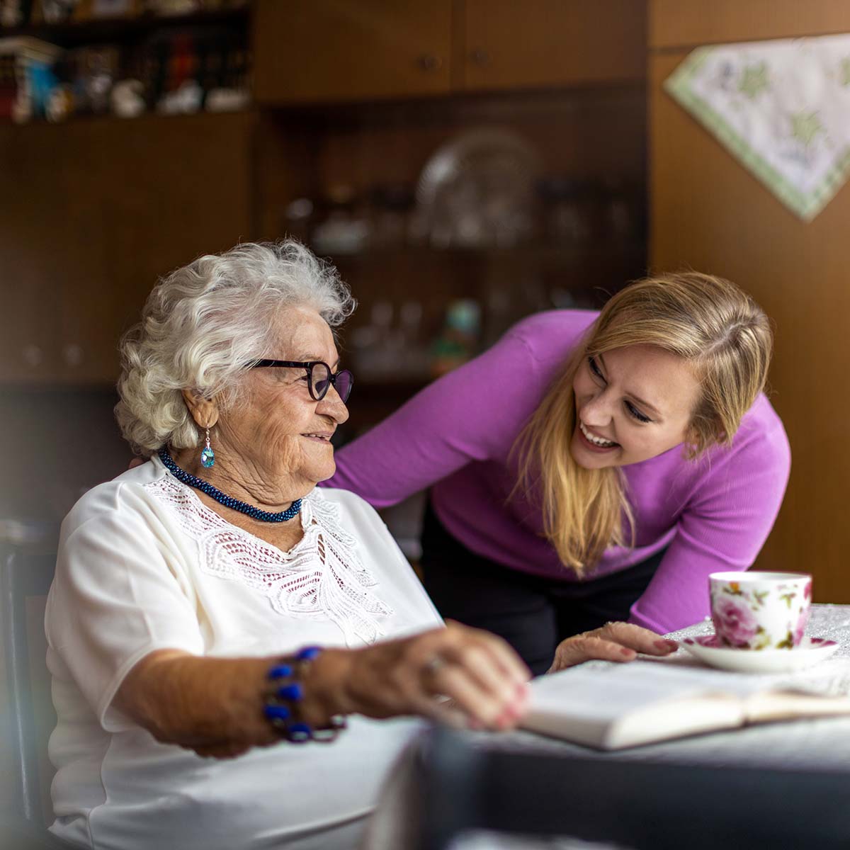 An adult child smiles and greets her mother as she's sitting at a table reading a book.