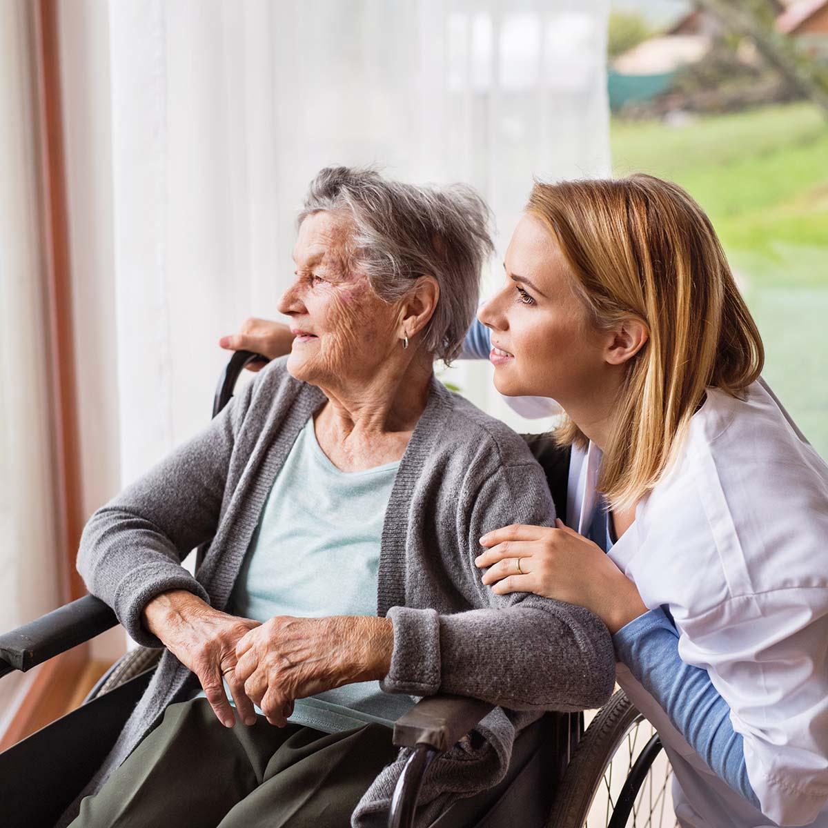 Nurse squatting next to a senior in a wheelchair looking out a window together.